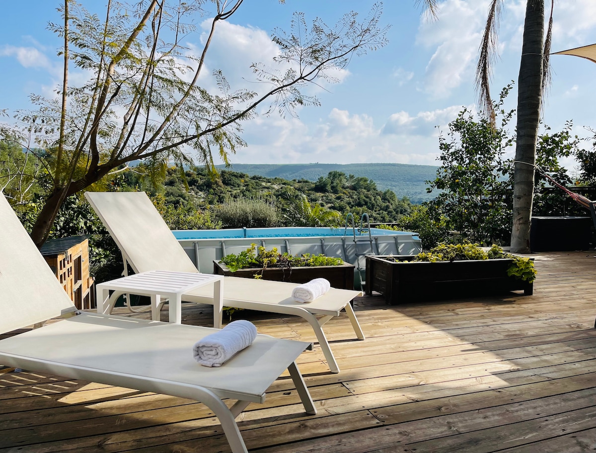 Two white lounge chairs with rolled towels are positioned on a wooden deck. A serene view of rolling hills and greenery is visible in the background, with an outdoor pool lined by plants. The sky features fluffy clouds and soft sunlight.