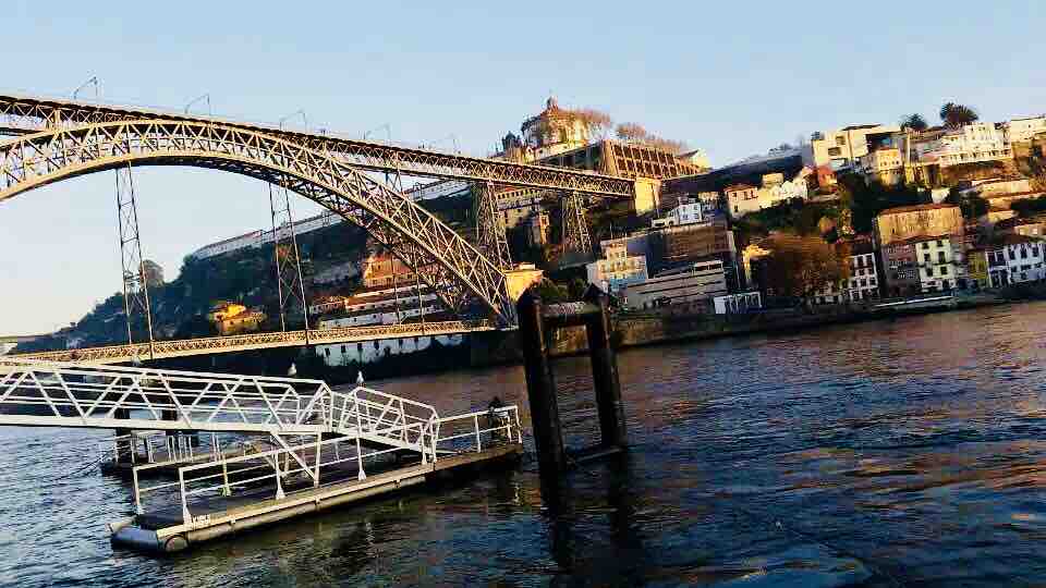 A striking view of a steel arch bridge spans over the river, connecting the two banks. A collection of colorful buildings is perched on the hillside, reflecting a mix of architectural styles. The water ripples gently, capturing the warm hues of the sunset.