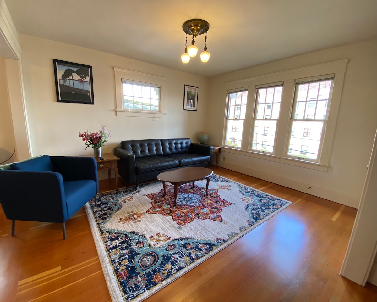 A spacious living area features a dark leather sofa and a cozy armchair, accented by a central wooden table. Natural light streams through large windows adorned with decorative panes, illuminating a colorful area rug that adds warmth to the restored wooden floors.
