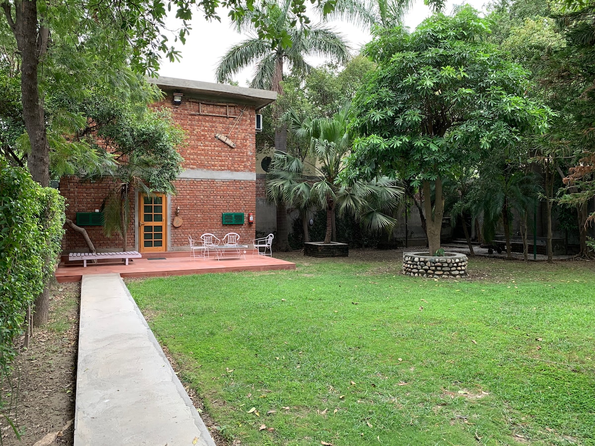 A spacious lawn is surrounded by trees and lush greenery, featuring a brick house with a welcoming entrance. A paved pathway leads to a patio area with outdoor seating, while a small circular stone feature sits within the garden, enhancing the natural ambiance.