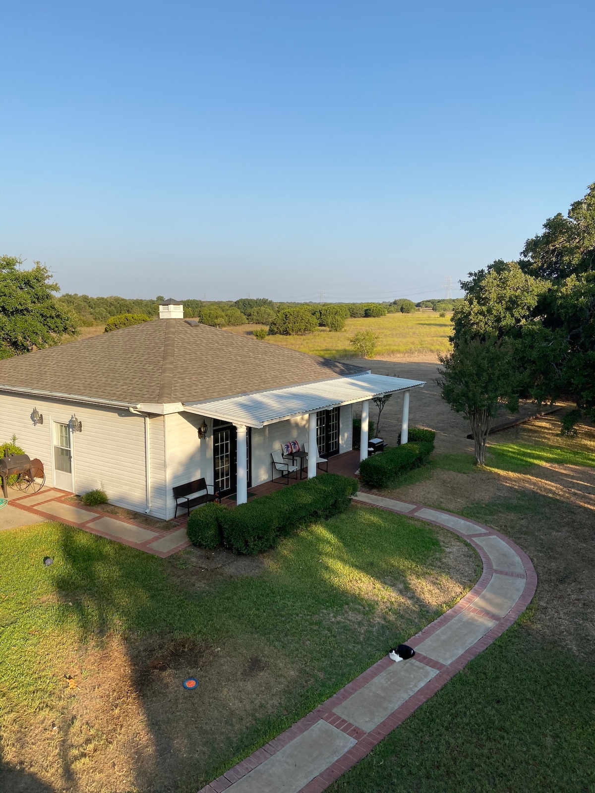 A charming cottage is framed by lush greenery, featuring a covered porch with seating. The winding pathway leads to the entrance, complemented by surrounding open fields and trees. The clear sky above creates a serene backdrop, enhancing the peaceful setting of the cottage.