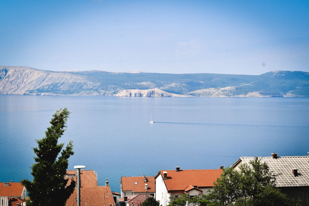 A panoramic view of the Kvarner Bay is captured, featuring calm blue waters and distant hills. The image shows a few rooftops of nearby houses with terracotta tiles, framed by greenery in the foreground. A small boat can be seen moving across the water.