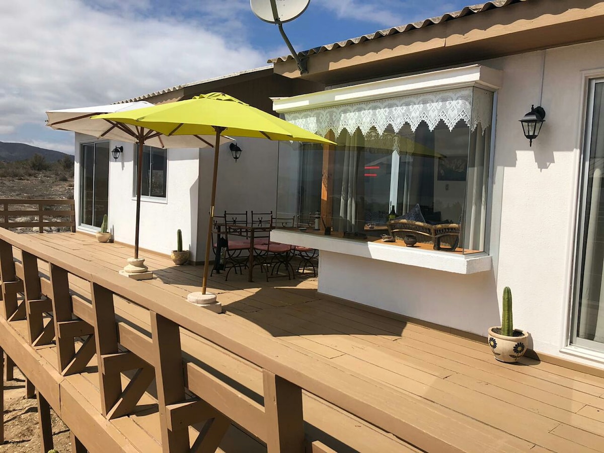 A wooden deck features several outdoor seating areas under large umbrellas. A spacious window allows for natural light to enter a nearby room, while decorative elements like potted plants add charm. The surrounding landscape shows a mix of desert terrain and distant hills.