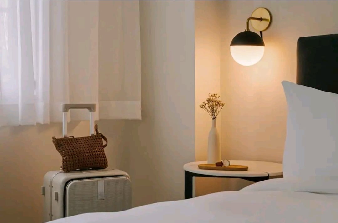 A bedside table is positioned next to a spacious bed, accompanied by a soft-glow wall lamp. A stylish suitcase is placed nearby, along with a small vase holding dried flowers, adding a subtle touch of decor.