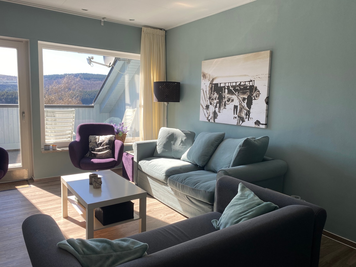A living room features a light blue wall and a large window offering a view of the surrounding nature. Two comfortable sofas face a minimalist coffee table. A piece of black and white artwork adorns the wall above the couch, adding a touch of style to the space.