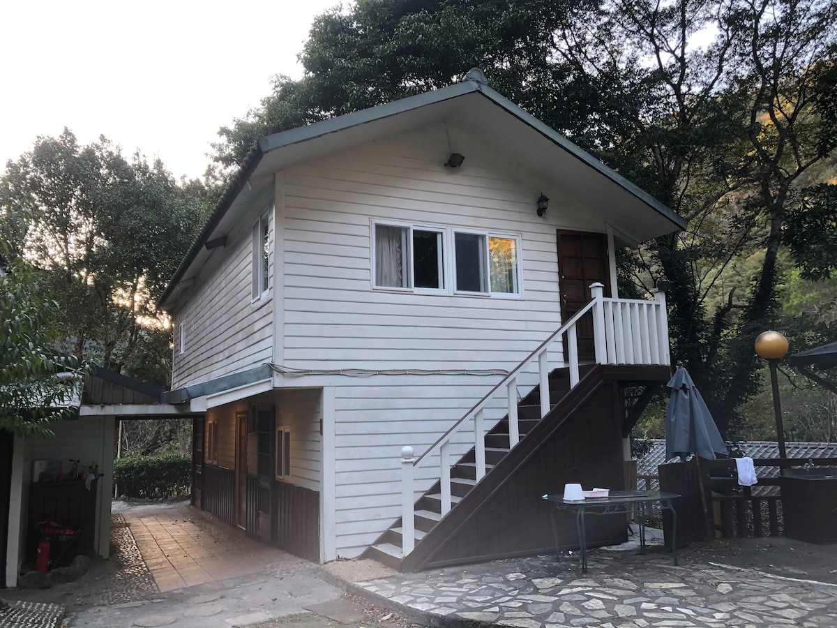 A two-story wooden building features a white exterior and a sloped roof. A set of stairs leads to the upper level, where large windows provide natural light. Surrounding greenery enhances the tranquil setting, and a stone pathway guides visitors to the entrance.