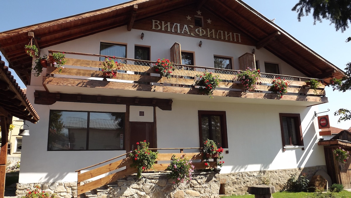 The two-story exterior of Villa Filip is presented, featuring a wooden balcony adorned with flowers. A welcoming entrance is highlighted by a wheelchair-accessible ramp, and large windows provide natural light to the interior. The combination of stone and wood adds character to the structure.
