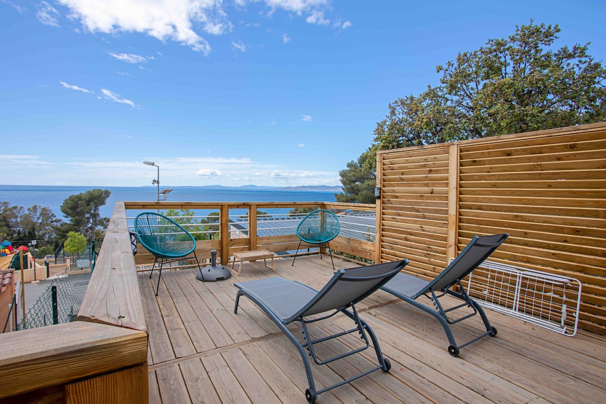 A spacious outdoor terrace is shown, featuring two lounge chairs positioned on a wooden deck. The unobstructed view of the coastline and sea extends beyond, complemented by the natural surroundings, which include trees and a glimpse of recreational areas.
