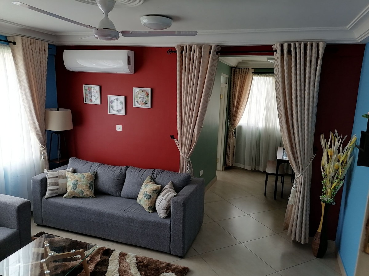 A living area is presented with a comfortable gray sofa adorned with decorative pillows. Light drapes frame large windows, allowing natural light to illuminate the space. A lamp stands beside the sofa, and nearby, a small desk area is visible in the adjoining room.