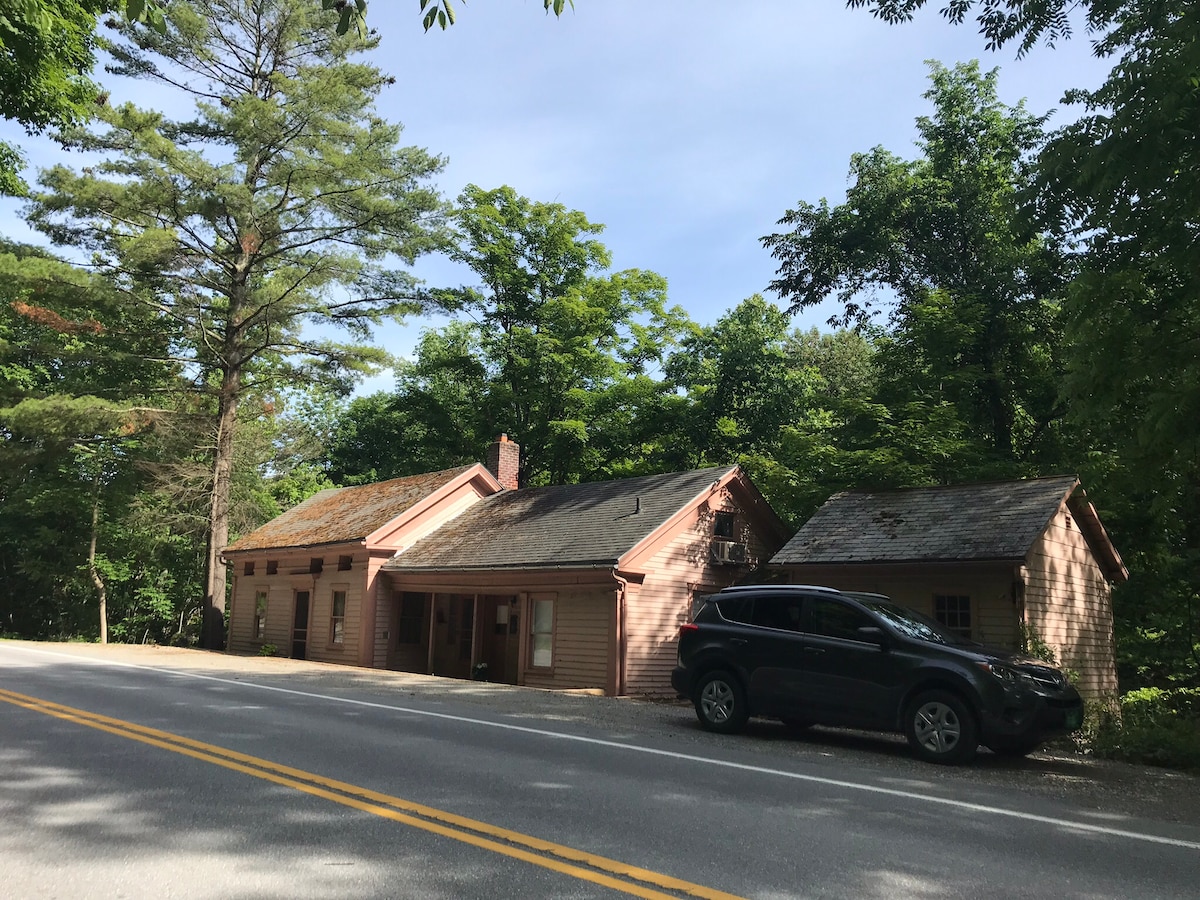 The Pink House, a historic structure, is set among lush greenery, showcasing its unique siding and charming architecture. A vehicle is parked along the roadside, which provides access to the house. Tall trees surround the property, contributing to a serene atmosphere.