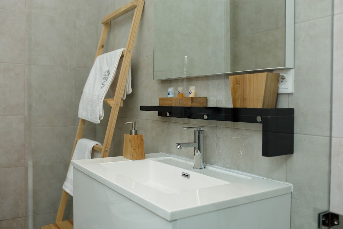 A modern bathroom features a sleek white sink with a minimalist design. Natural wood accents are highlighted in the soap dispenser and storage. Soft, folded towels rest on a wooden ladder. A large mirror is mounted above the sink, enhancing the spacious feel.