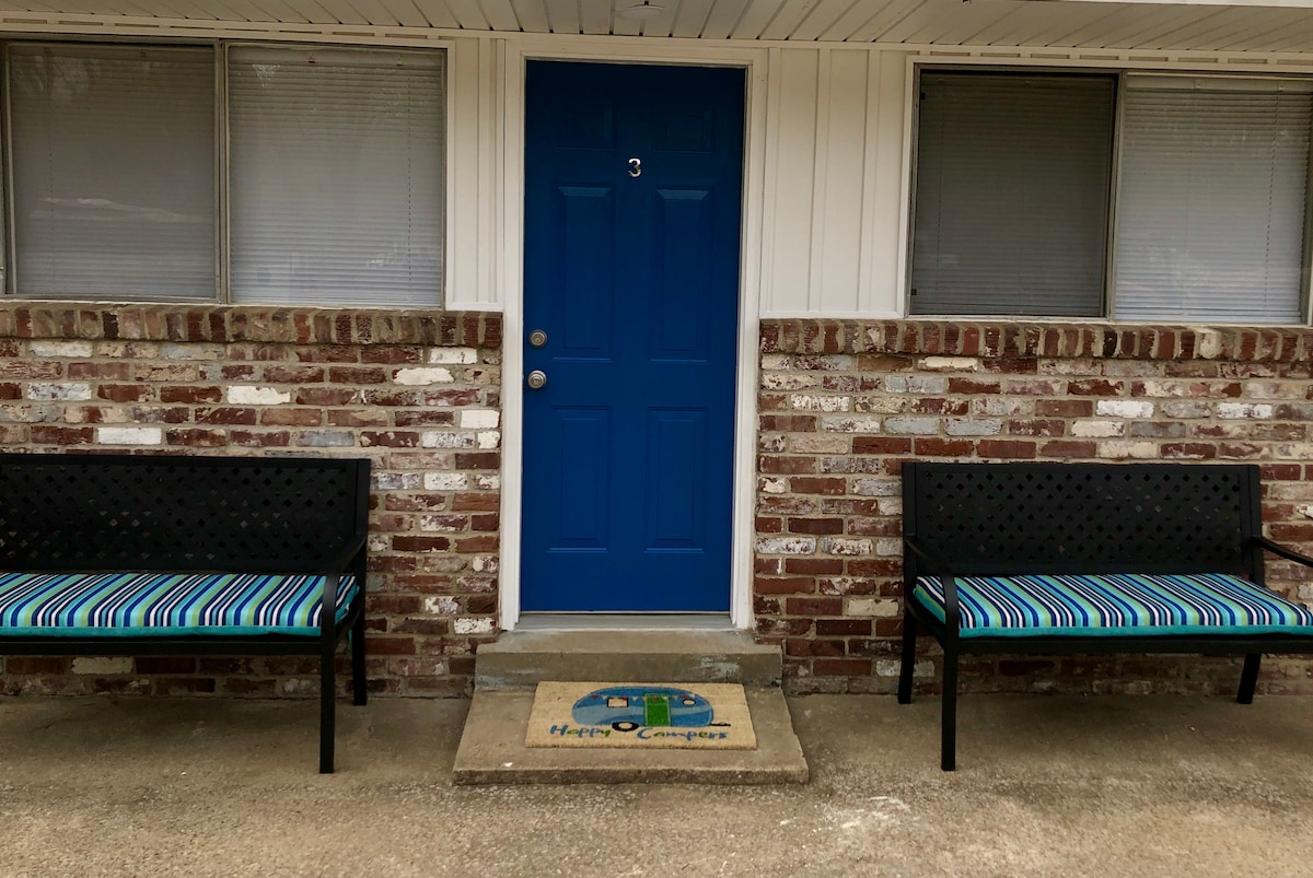A welcoming entrance displays a bright blue door, flanked by two black benches with striped cushions. A doormat featuring a cartoon camper adds a playful touch. Light filters through the two windows on each side, illuminating the brick exterior.