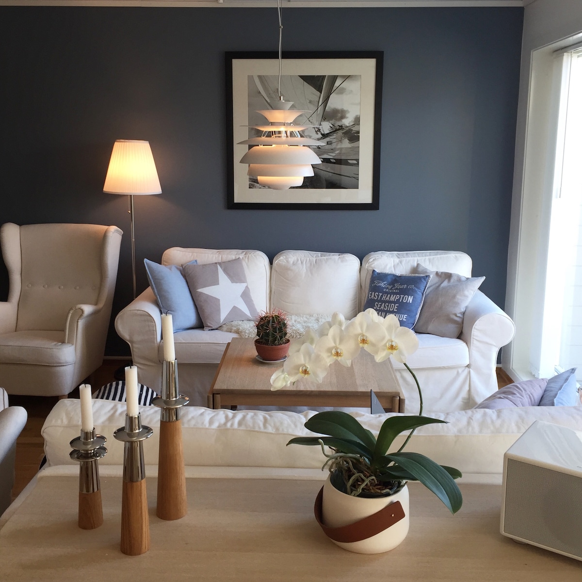 A cozy living area displays a light-colored sofa adorned with various decorative pillows. Two armchairs are positioned nearby, and a wooden coffee table sits at the center. A potted orchid adds a touch of greenery, while a softly glowing lamp illuminates the space against a dark blue wall.