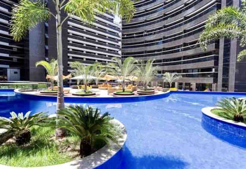 A large swimming pool is surrounded by lush greenery and palm trees, with sun loungers placed on the deck. The building's modern architecture is reflected in the water, contributing to a serene outdoor atmosphere.