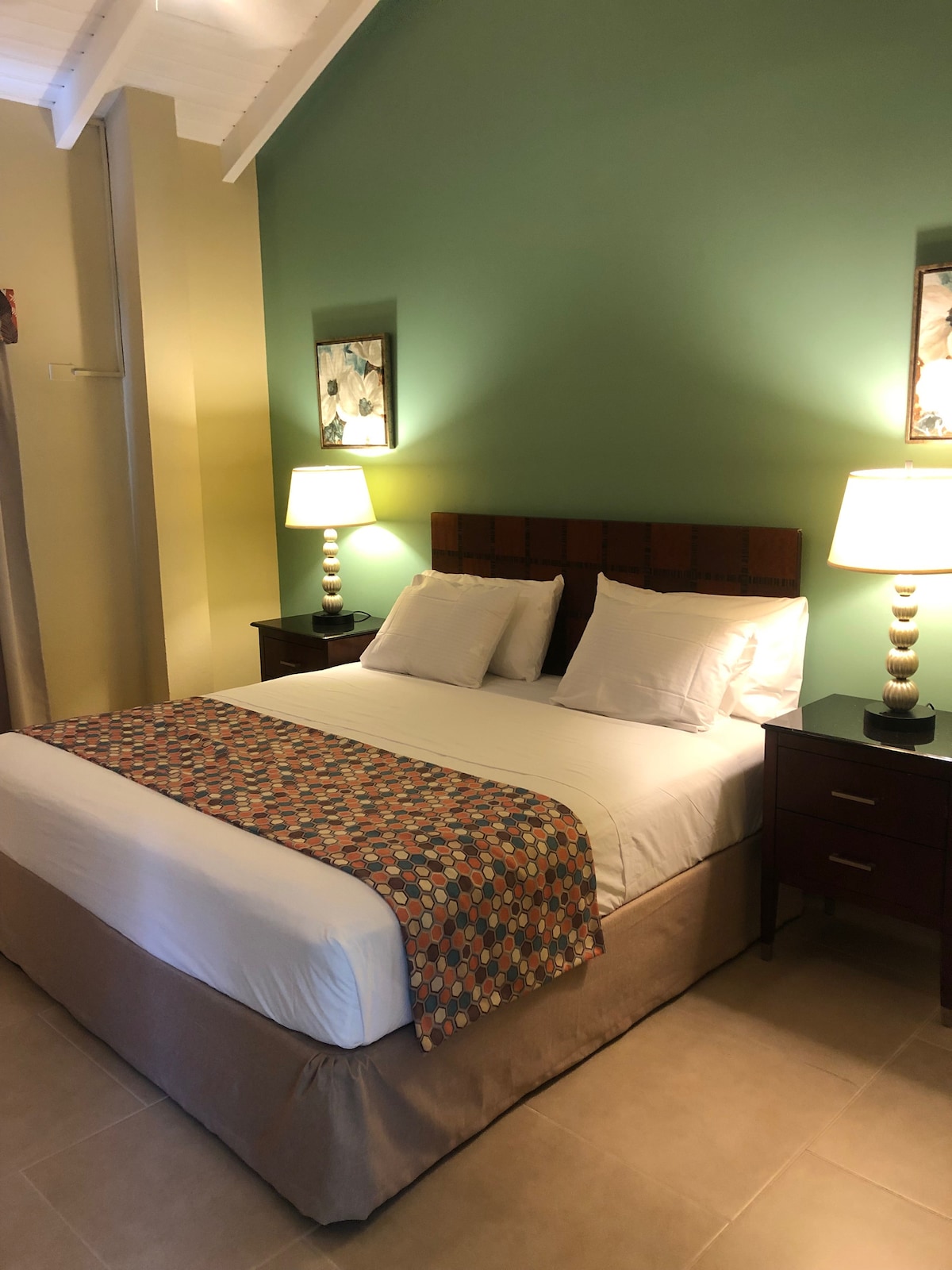 A tastefully decorated room features a spacious bed with crisp white linens and a colorful throw. Nightstands with lamps are positioned on either side of the bed, and artwork is displayed on the green accent wall, providing a calming environment.