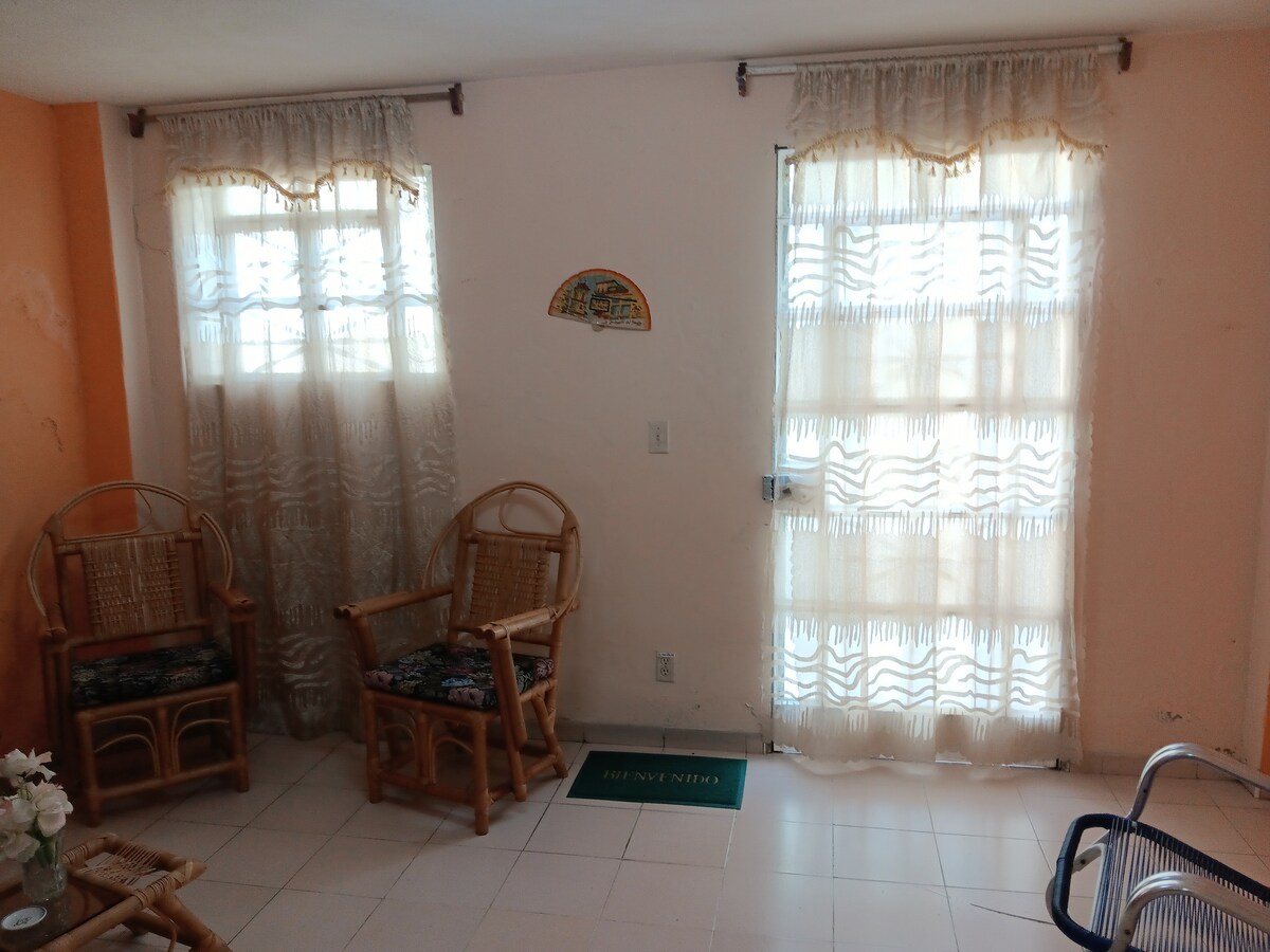 A light-filled seating area features two bamboo chairs with colorful cushions, positioned beside a window with sheer curtains. The wall displays a decorative item, and a door with a glass panel leads to the outside, creating a welcoming space for relaxation.