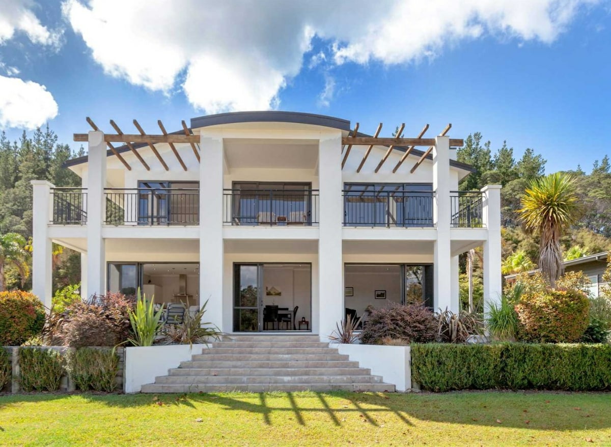 A modern two-story house is depicted with a large entrance stairway framed by shrubs. Generous balconies overlook the garden, and expansive glass doors provide views of the interior. The building is surrounded by palm trees and lush greenery against a backdrop of blue skies and scattered clouds.
