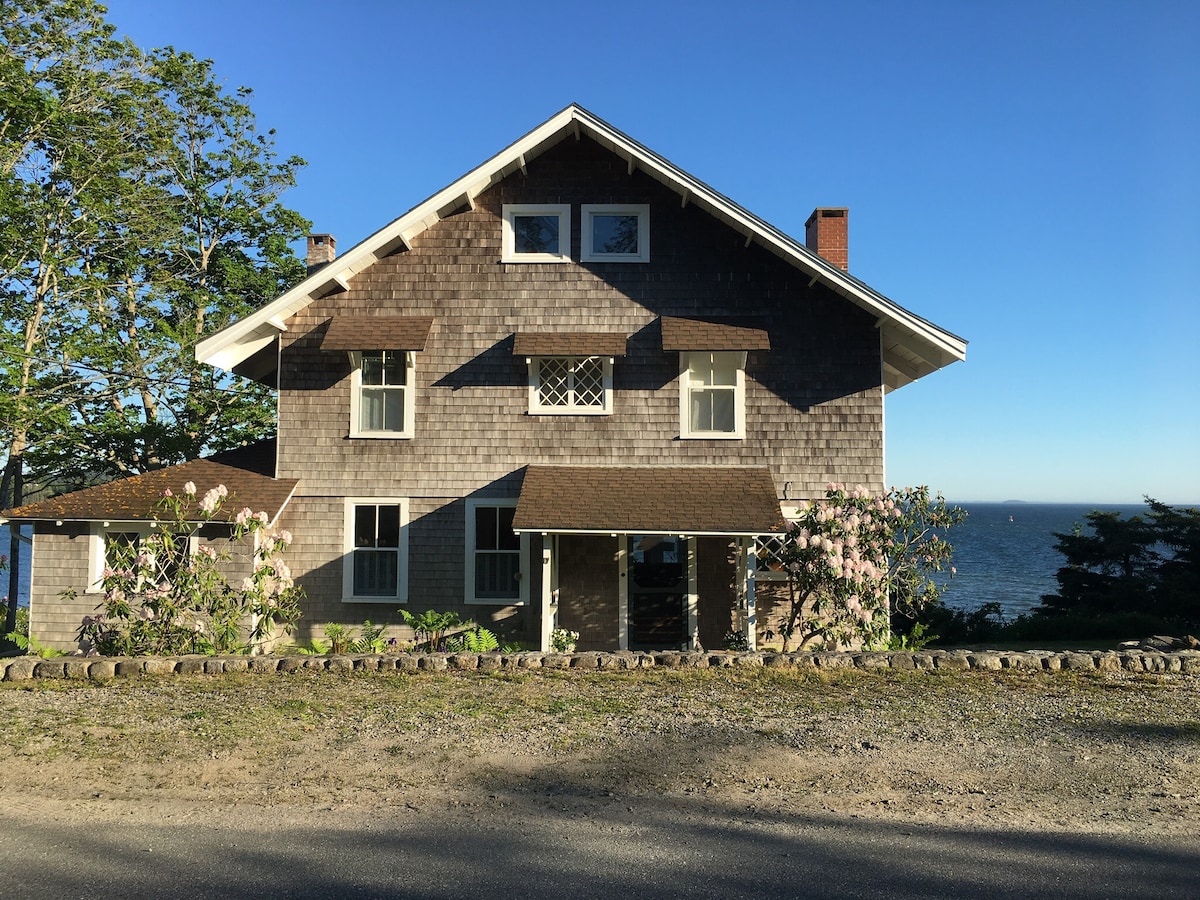 The exterior of Shore Cottage is displayed, featuring rustic wooden shingles and multiple windows. The front porch is supported by stone pillars, with flowering plants lining the entrance. A clear view of the water is visible in the background, under a bright blue sky.