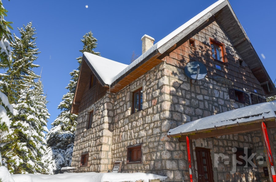 A multi-story building features a charming stone exterior, complemented by wooden accents. Snow-covered trees surround the house, and bright blue skies are visible above, creating a serene winter landscape.