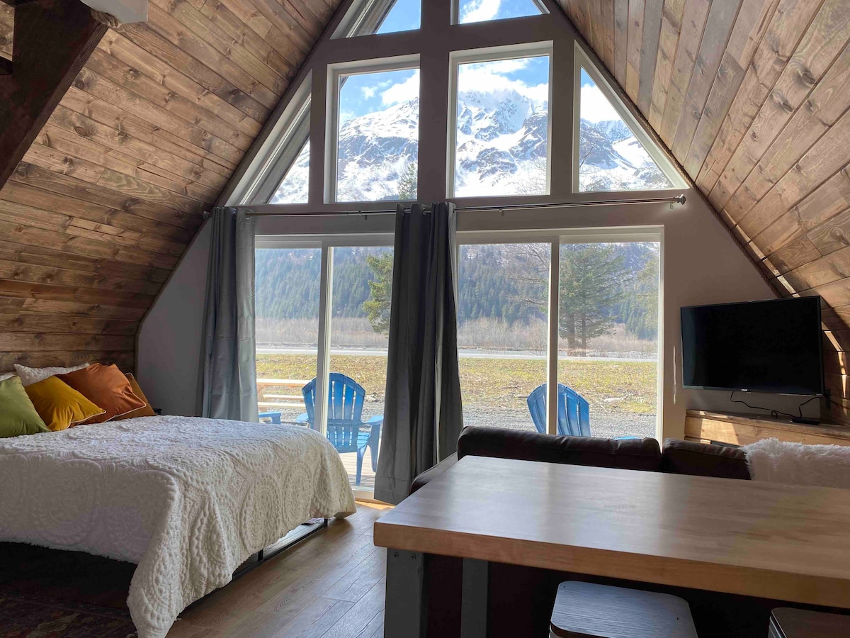 Large windows frame stunning mountain views, allowing natural light to fill the space. A cozy sleeping area features a spacious bed with a textured white coverlet. Soft throw pillows in warm colors add comfort, complemented by a small dining table and lounge chairs on the patio.