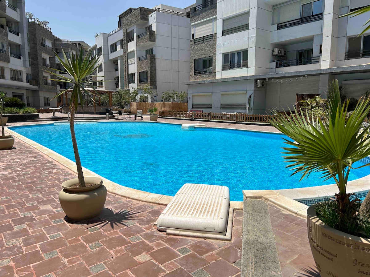A crystal-clear swimming pool is framed by textured stone and lush greenery, with a sunken lounge area and sunbeds nearby. The pool's vibrant blue water is contrasted by the light-colored tiles surrounding it, enhancing the serene ambiance of the gated community.