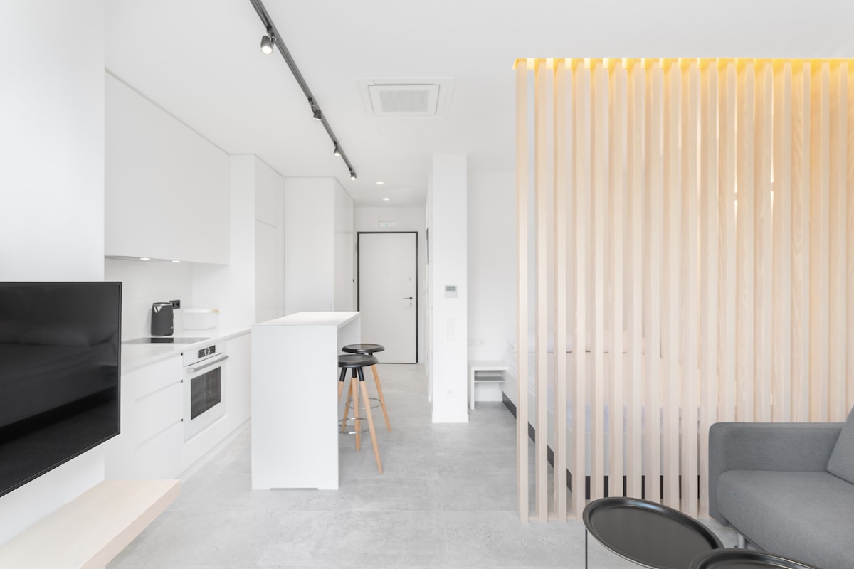 A modern studio apartment features a minimalist design with a neutral color palette. A kitchen area includes sleek cabinetry and modern appliances. A wooden partition provides a subtle separation between the living space and the sleeping area, while natural light illuminates the room.