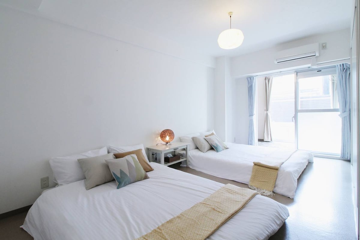 The room features two spacious beds, dressed in crisp white linens. A light fixture hangs from the ceiling, and natural light enters through the large window, which is framed by light blue curtains, creating a calm and inviting space.