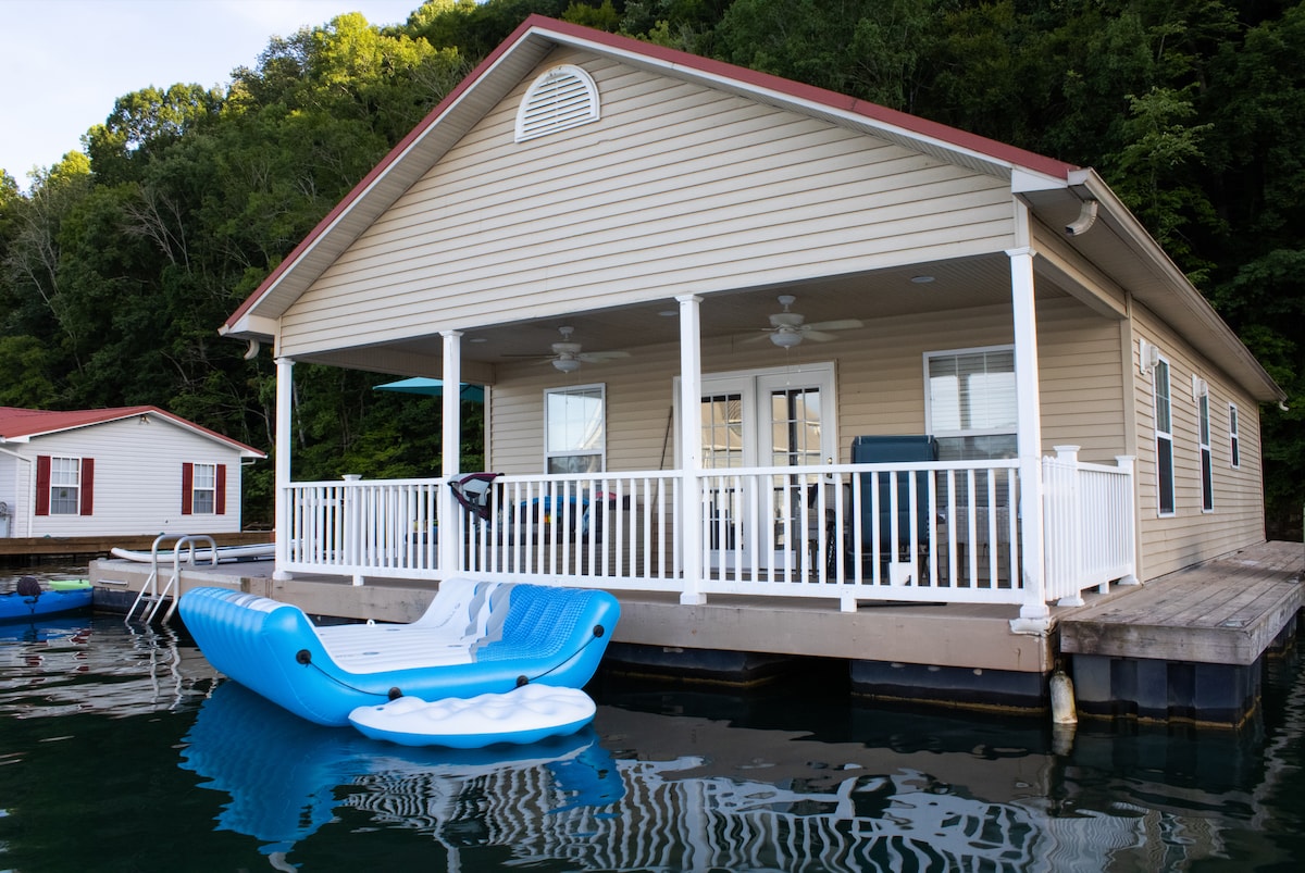 The floating lake house features a spacious porch with ceiling fans and comfortable seating areas. A blue paddle boat is docked nearby, complementing the serene water setting. Lush green trees rise behind the house, enhancing the natural ambiance of the location.