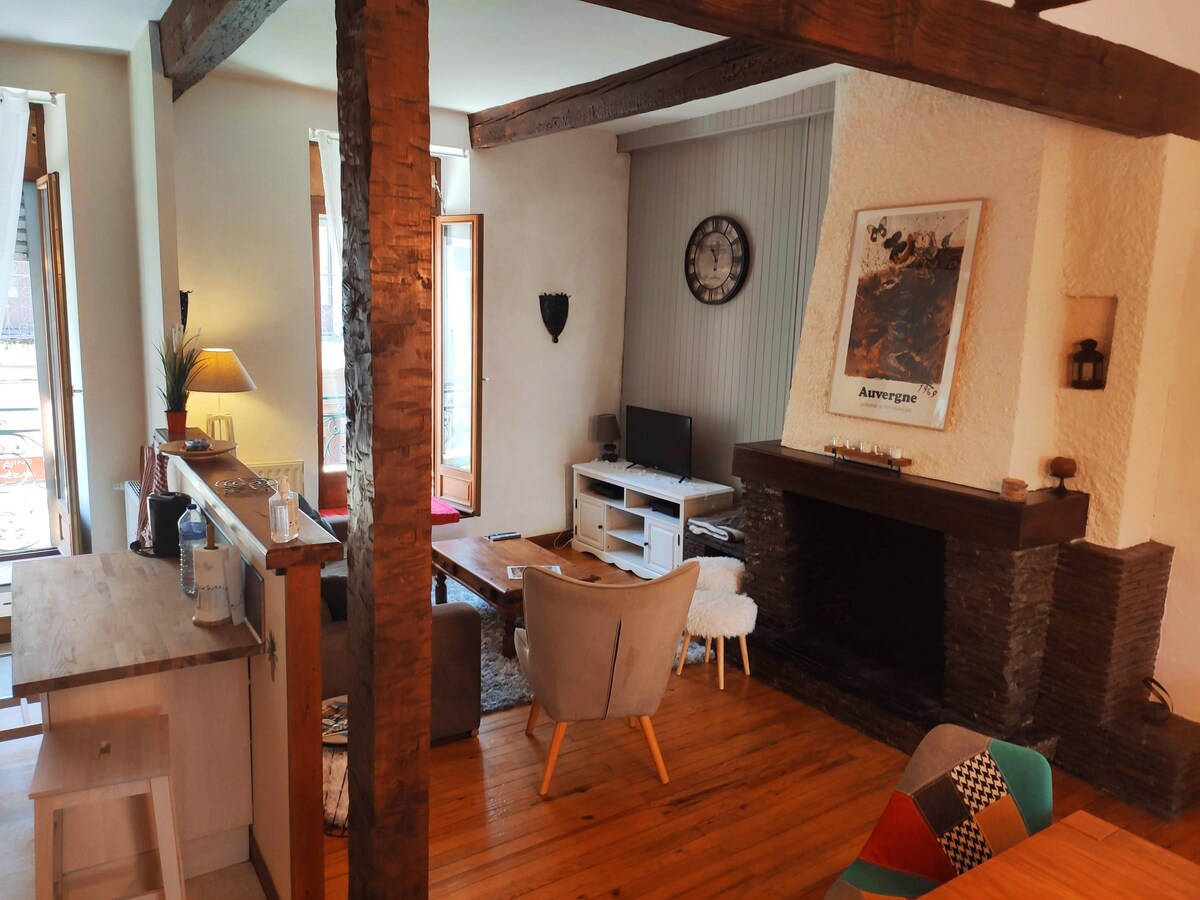 A cozy living area features a stone fireplace and a flat-screen TV. Light wood accents and natural light from large windows create a warm ambiance. A central coffee table accompanied by soft seating invites relaxation, while a decorative clock adds a charming touch to the space.
