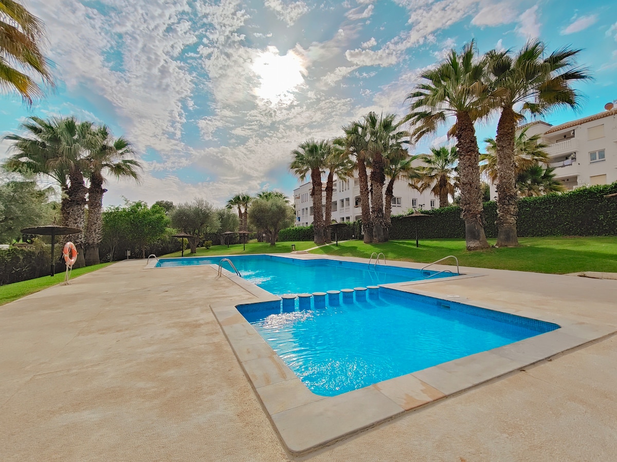 A communal pool surrounded by landscaped gardens features clear blue water under a bright sky. Palm trees line the pool area, offering a tropical ambiance. Lounge chairs are positioned along the poolside, providing ample space for relaxation.