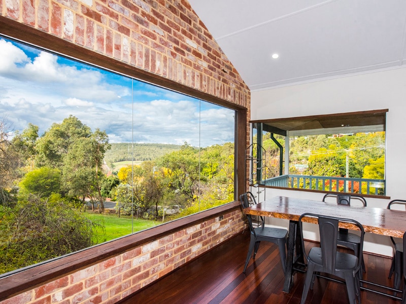 A large window showcases views of lush greenery and rolling hills. The dining area features a natural wood table complemented by black metal chairs. The warm tones of the brick walls and polished timber floors create a welcoming atmosphere. Natural light brightens the open space.