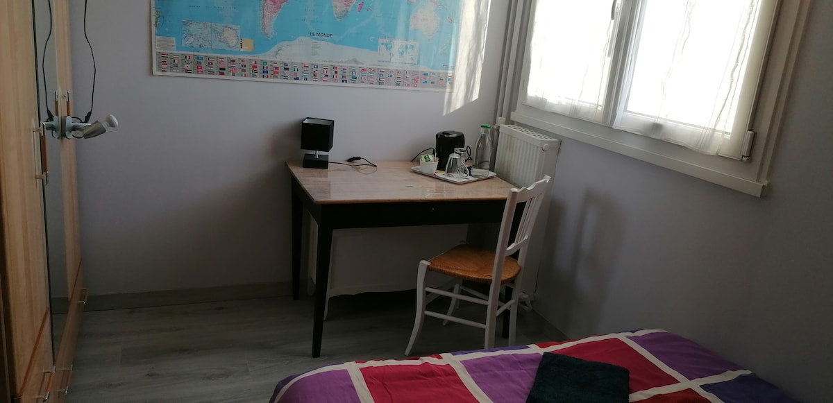 A bedroom is equipped with a desk facing a window, which allows natural light to fill the space. A map adorns the wall above the desk, alongside a small coffee station. A spacious bed is visible, covered with colorful bedding, and a chair is placed beside the desk.