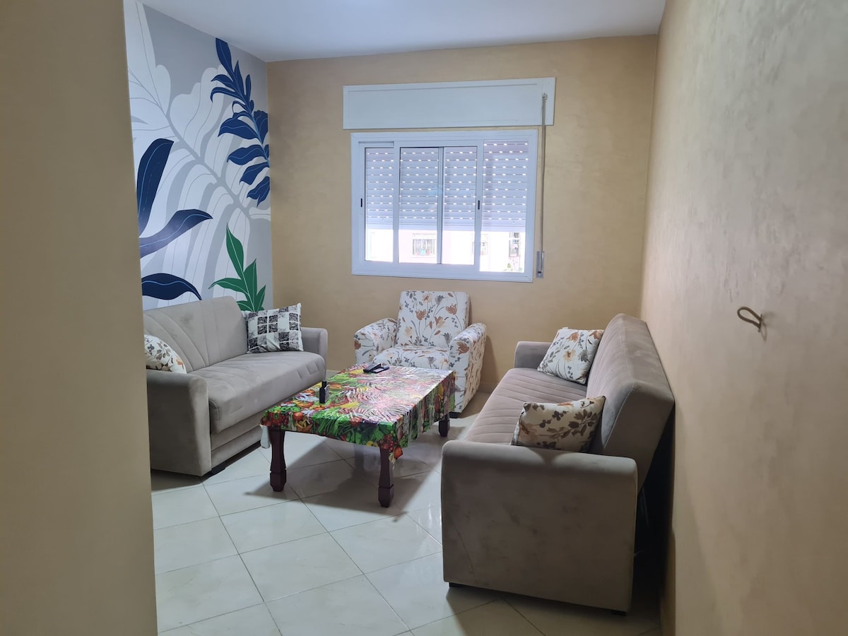 A cozy living area features light-colored sofas arranged around a vibrant, floral-patterned table. Natural light enters through a large window, adorned with textured shades. A mural of tropical leaves on one wall adds a touch of color to the neutral-toned space.
