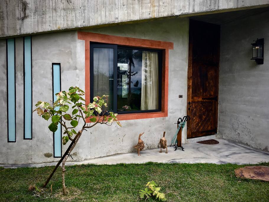 房門口,赭紅框住大窗、細長玻璃以黑線條修飾,斑葉黃槿種植於前方