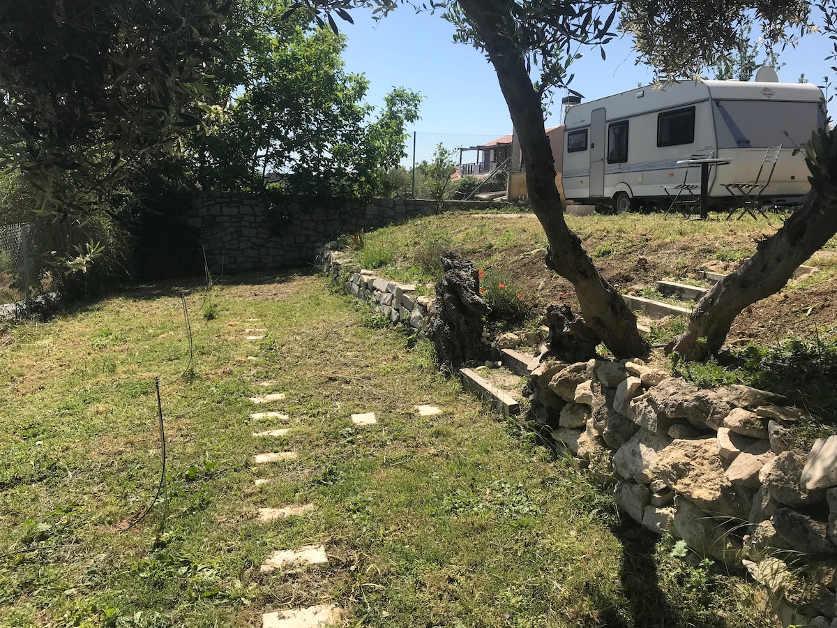 A well-maintained private garden is visible, featuring a grassy area with stepping stones leading towards a caravan. Olive trees provide shade, while nearby, a staircase ascends to a higher garden level. The vibrant greenery and open sky create a serene outdoor environment.