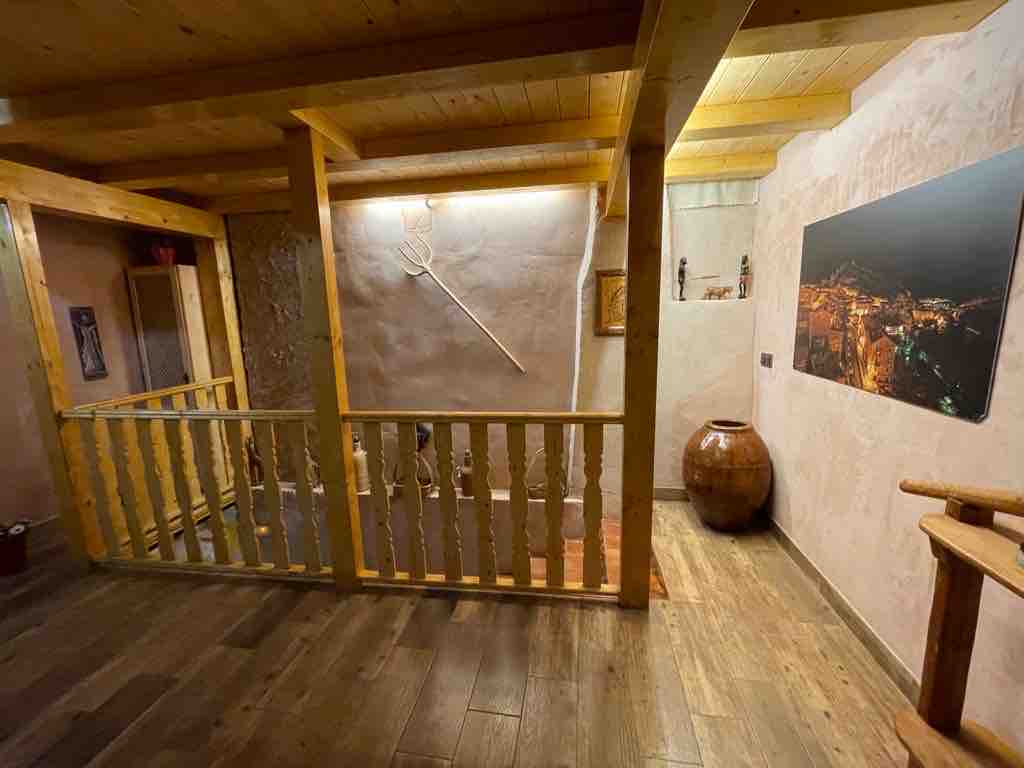 A warm wooden balcony overlooks a spacious area, featuring rustic decor highlighted by a large earthen vase and a decorative wall piece. Soft lighting enhances the natural materials used throughout, contributing to a cozy atmosphere in the common area.