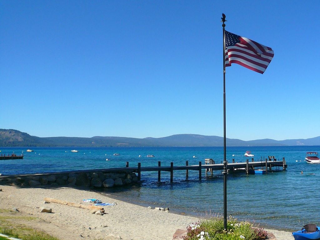 A view of a serene beach scene features a flag gently waving in the breeze near the shore. Clear blue waters reflect the sky, while a wooden pier extends into the lake. Distant mountains complete the picturesque landscape under a bright blue sky.