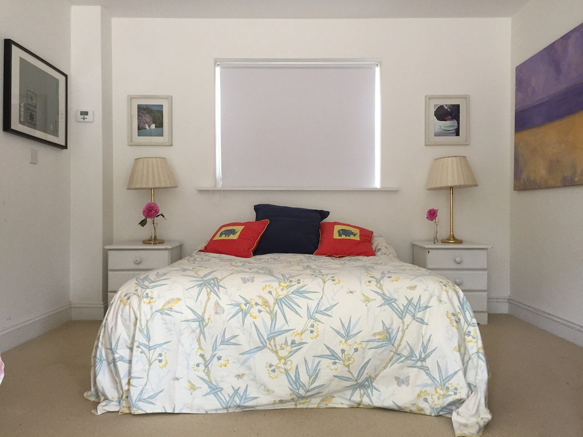 A spacious bedroom features a large bed adorned with a floral-patterned duvet and vibrant cushions. Nightstands on either side are topped with matching lamps. Artwork is displayed on the walls, and natural light enters through a window covered with a white shade.
