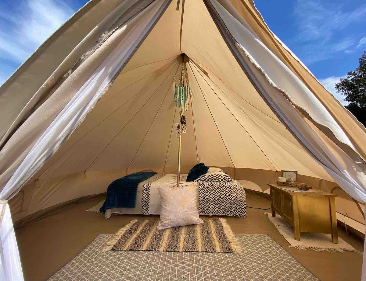 An inviting glamping tent is adorned with soft bedding, a decorative throw, and cushions. A small gold side table is positioned beside the bed, with a textured rug laid on the floor. Natural light enters through the tent's open flaps, creating a warm atmosphere.