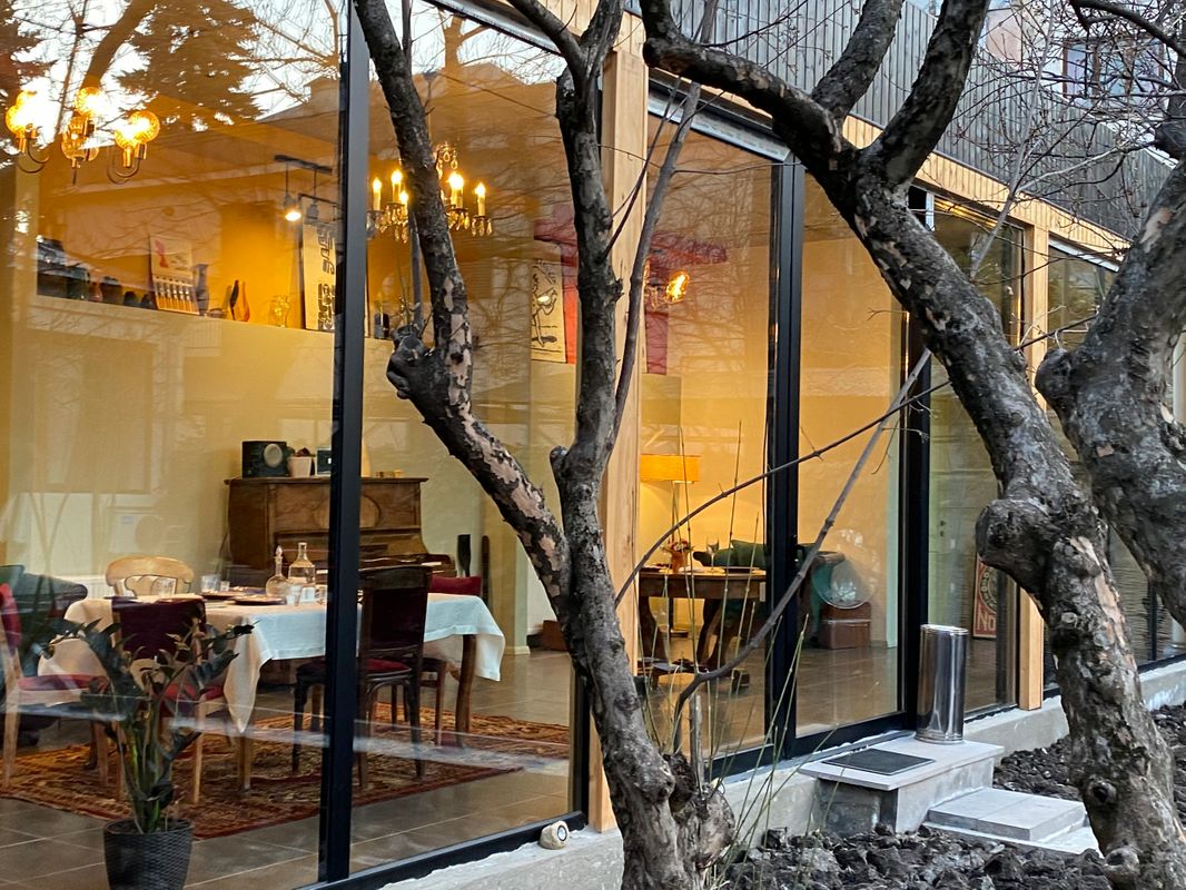 An exterior view showcases a vibrant dining space featuring large glass windows, allowing for natural light to flood the area. Inside, a dining table is elegantly set, accompanied by various chairs. A warm glow emanates from a chandelier, enhancing the inviting atmosphere.
