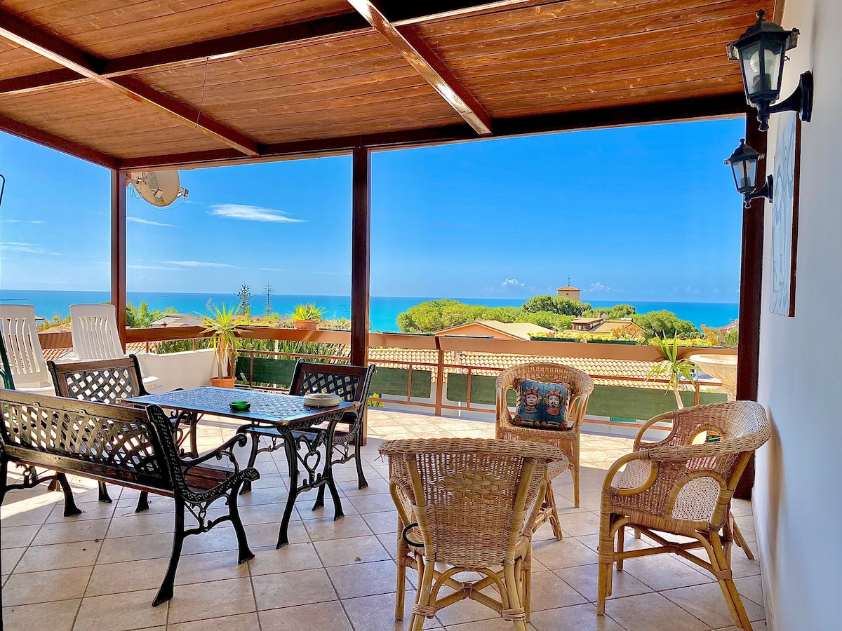 An outdoor terrace is captured, featuring a table surrounded by wicker and metal chairs. The sea and coastline are visible in the background, framed by greenery. The space is covered with a wooden pergola and is enhanced by clear blue skies.