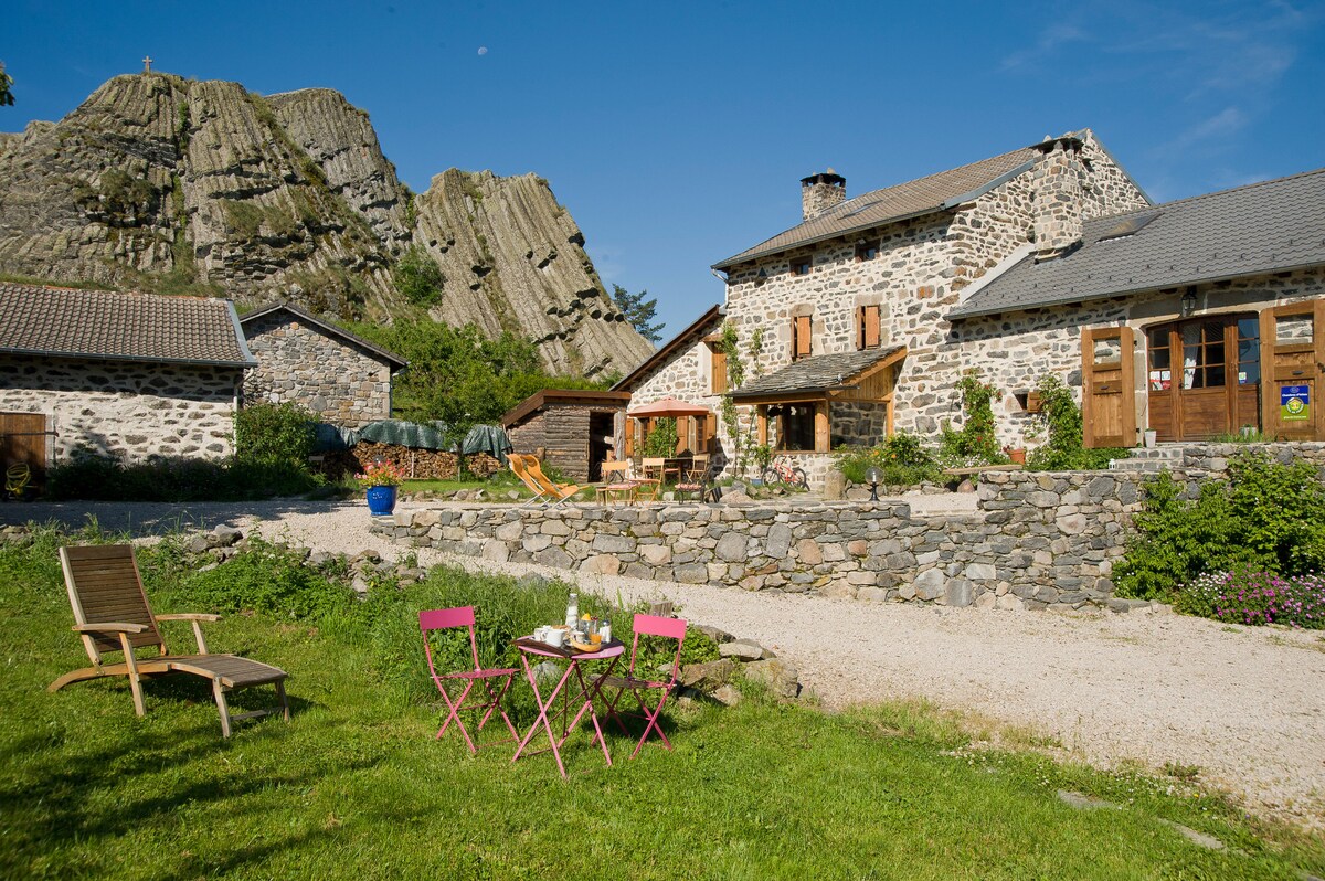 A rustic stone cottage is shown, with the dramatic backdrop of rocky cliffs. In the foreground, a small table with two pink chairs is set up on green grass, inviting calm moments outdoors. The sun illuminates the inviting facade of the cottage against a clear blue sky.