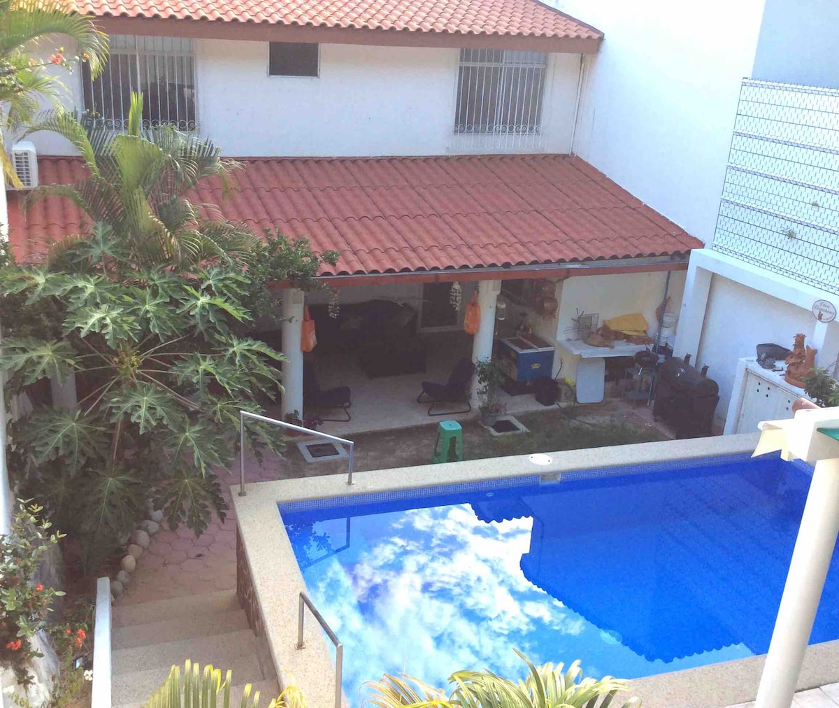 A private pool is surrounded by a spacious outdoor area featuring comfortable seating and lush greenery. A covered patio offers shade, with elements visible such as a kitchenette and lounge chairs. The pool reflects the sky and surrounding foliage, creating a serene outdoor retreat.