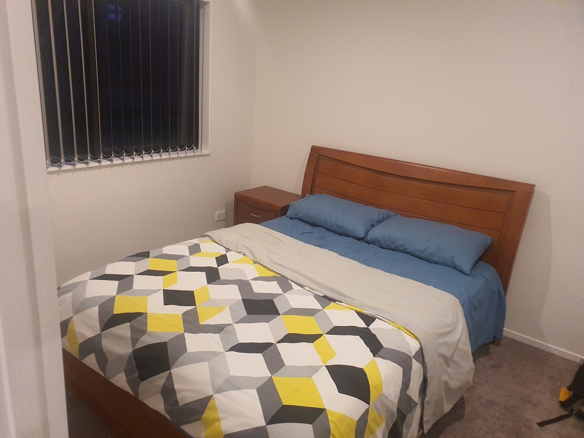 A spacious bedroom is equipped with a large wooden bed featuring a patterned quilt in grey, yellow, and white. Light blue pillows complement the bedding. A tall window with vertical blinds allows for natural light, alongside a bedside table positioned on one side.