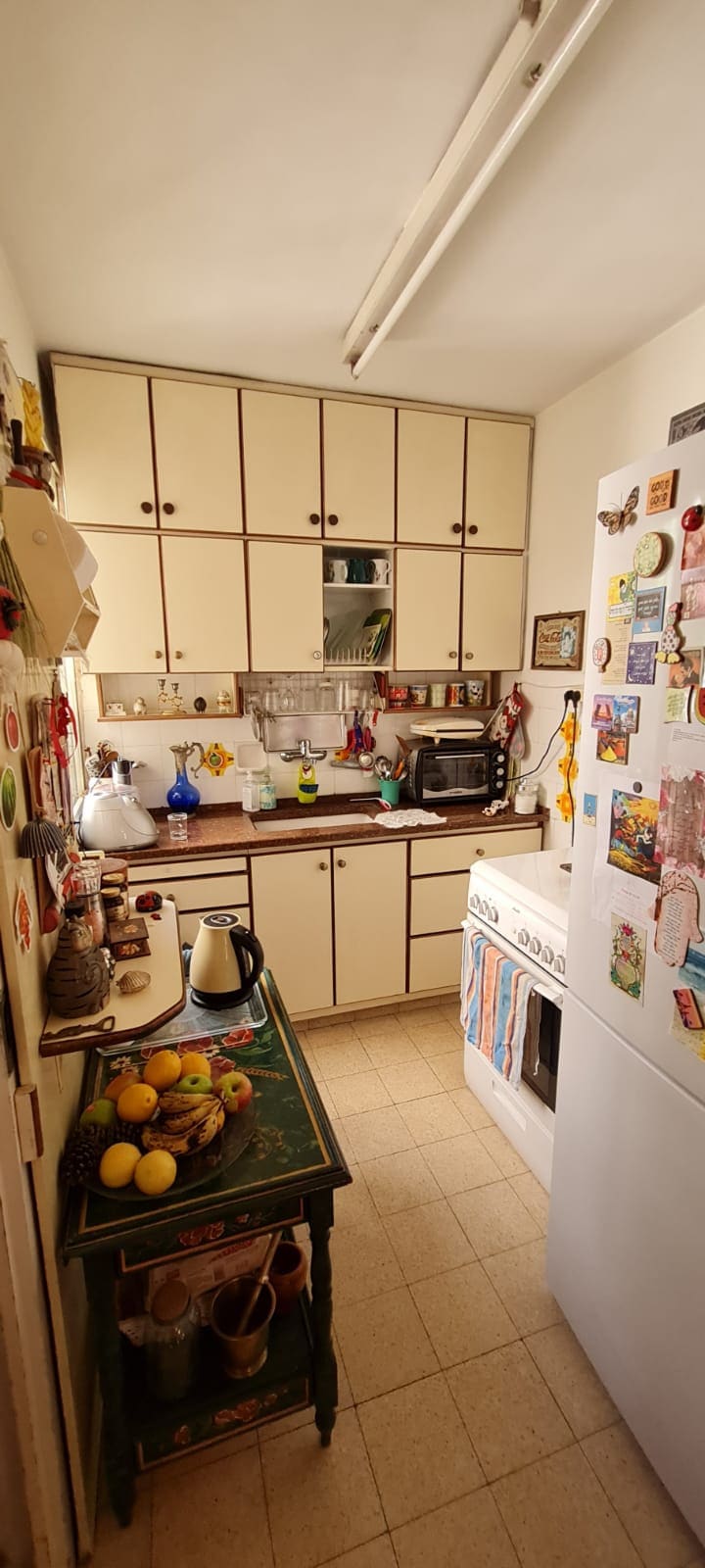 A compact kitchen is equipped with a white refrigerator and a countertop oven. Wooden cabinets line the walls, offering ample storage. A collection of colorful magnets and notes adorns the fridge. A table displaying fresh fruits adds a touch of home to the space.