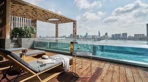 A sunlit terrace features a sleek outdoor lounge area with two sun loungers and a small table. A panoramic view of the city skyline is visible in the background, while a glistening infinity pool extends beyond the edge of the terrace.