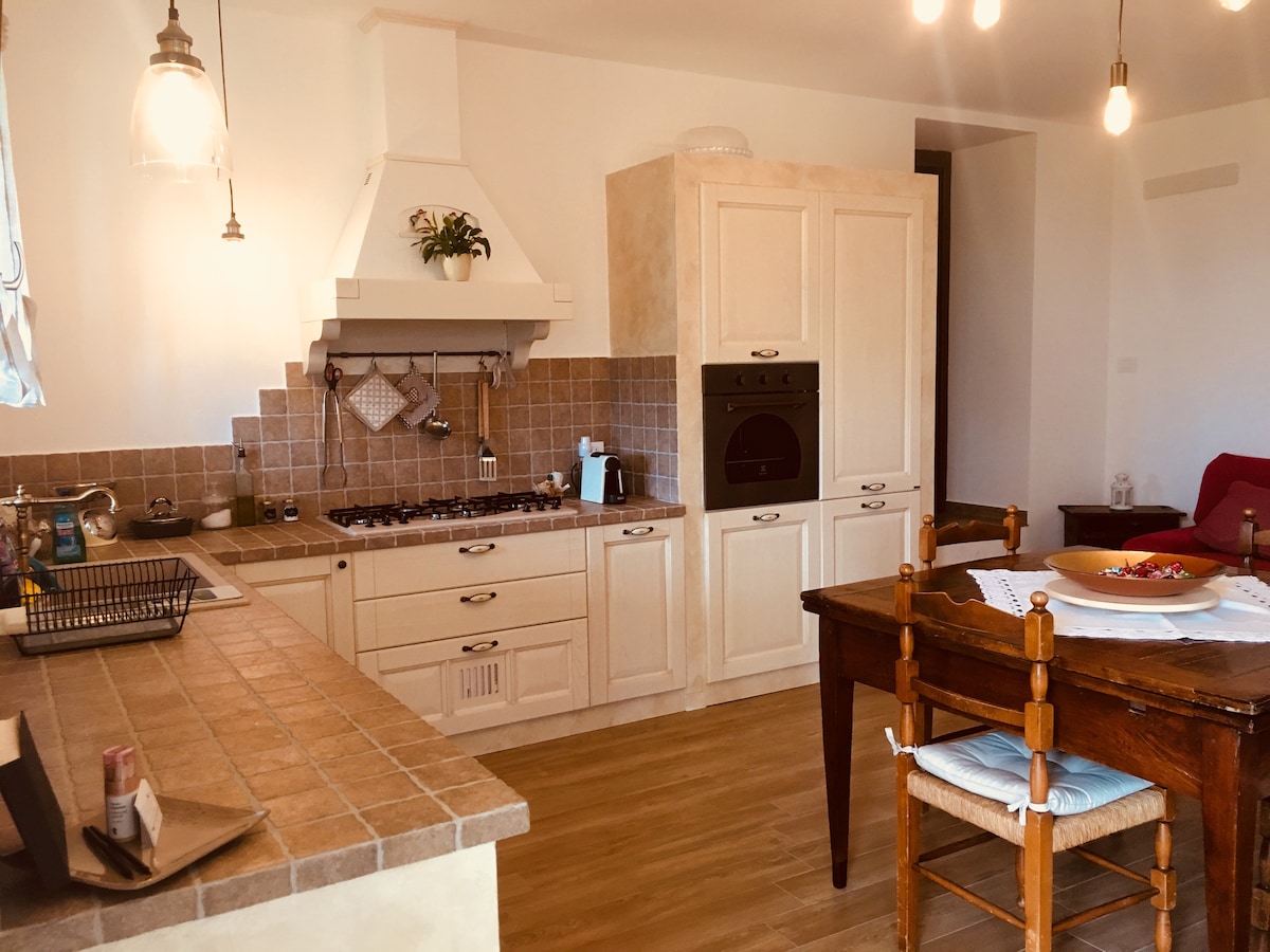 A cozy kitchen area is seen featuring wooden cabinetry and a rustic dining table with chairs. Natural light fills the space, highlighting the stove and oven built into the cabinetry. A kettle and various kitchen items are arranged on the countertops.
