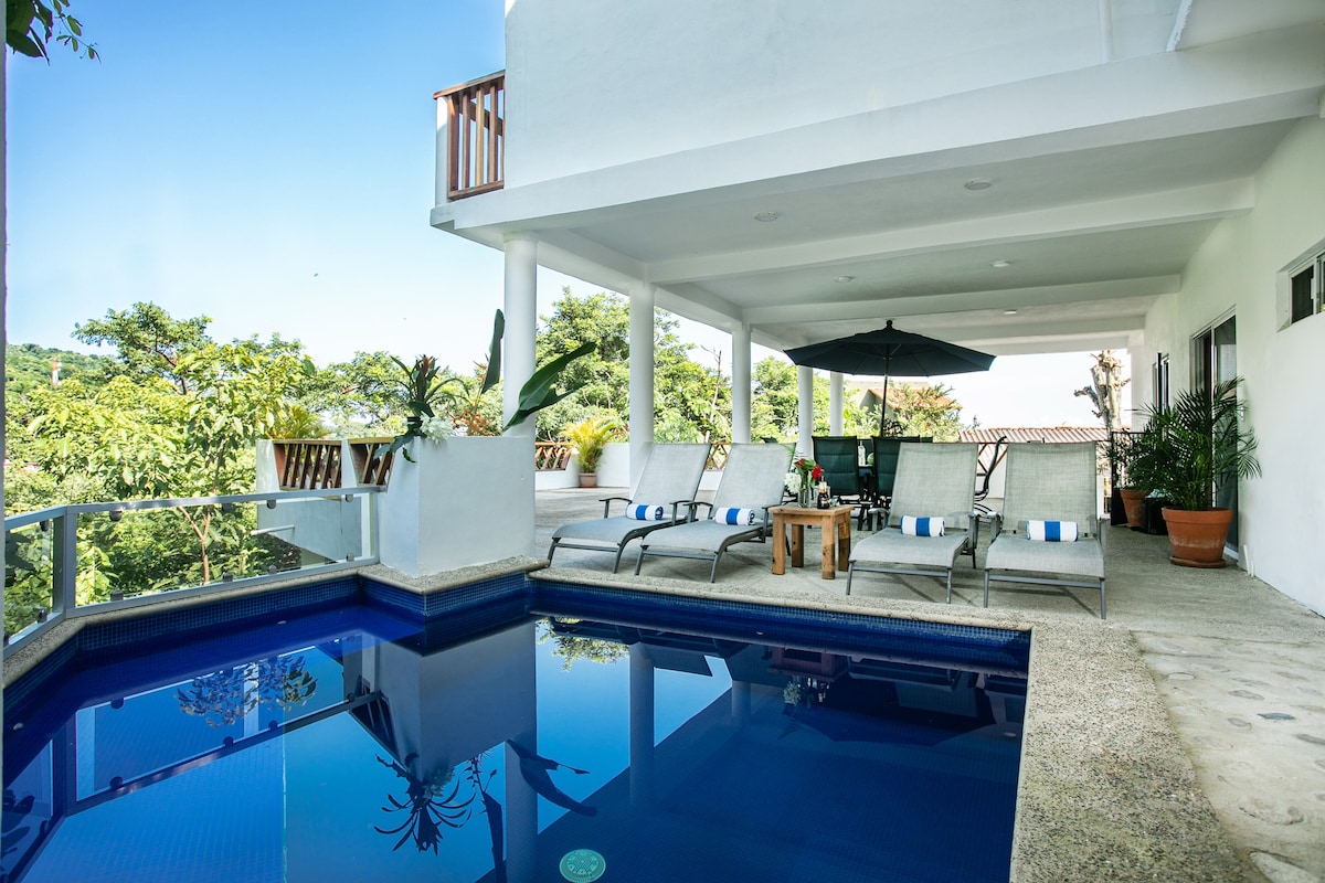 An outdoor patio overlooks a private pool, surrounded by lounge chairs and potted plants. An umbrella provides shade for seating areas, while lush greenery frames the view, creating a serene setting for relaxation.