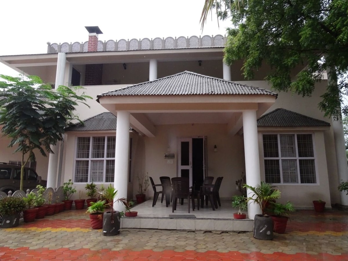 The front view of the spacious farm stay is showcased, featuring a covered porch with outdoor seating. Potted plants line the entrance, and large windows provide ample natural light. The roof is adorned with a decorative trim, complementing the white columns and tiled floor.