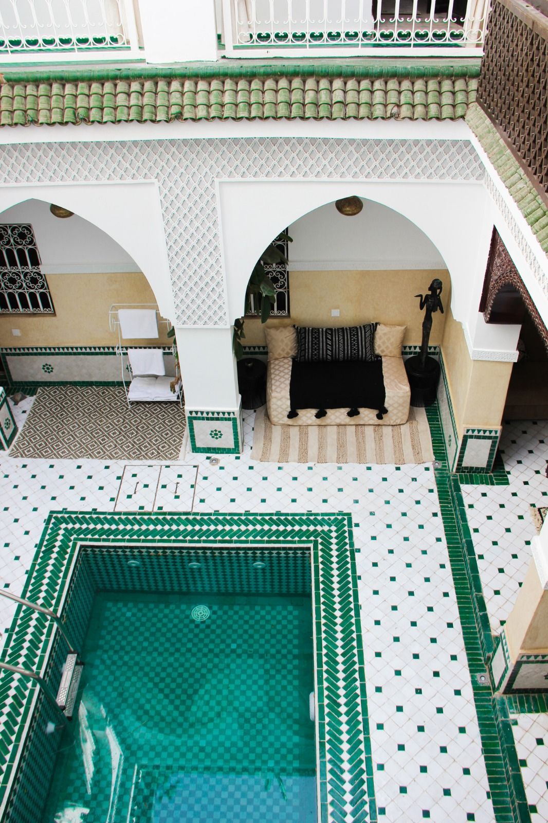 An inviting courtyard features a serene pool surrounded by intricate green and white tiles. Arched doorways lead to comfortable seating areas, providing spaces for relaxation. The layout reflects traditional Moroccan design, enhancing the ambiance with a sense of tranquility.
